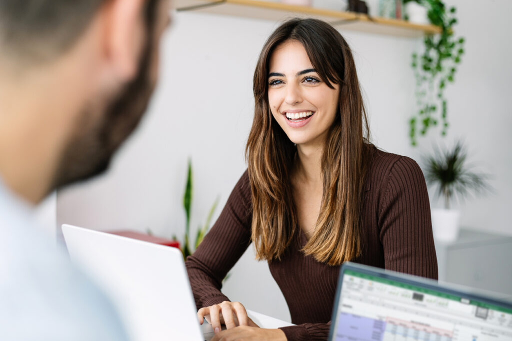 Smiling young coworkers working together with laptop in the office. Pretty millennial businesswoman talking to colleague at co-working workplace. Young couple telecommuting at home. Entrepreneur people, startup and teamwork and home office concept.