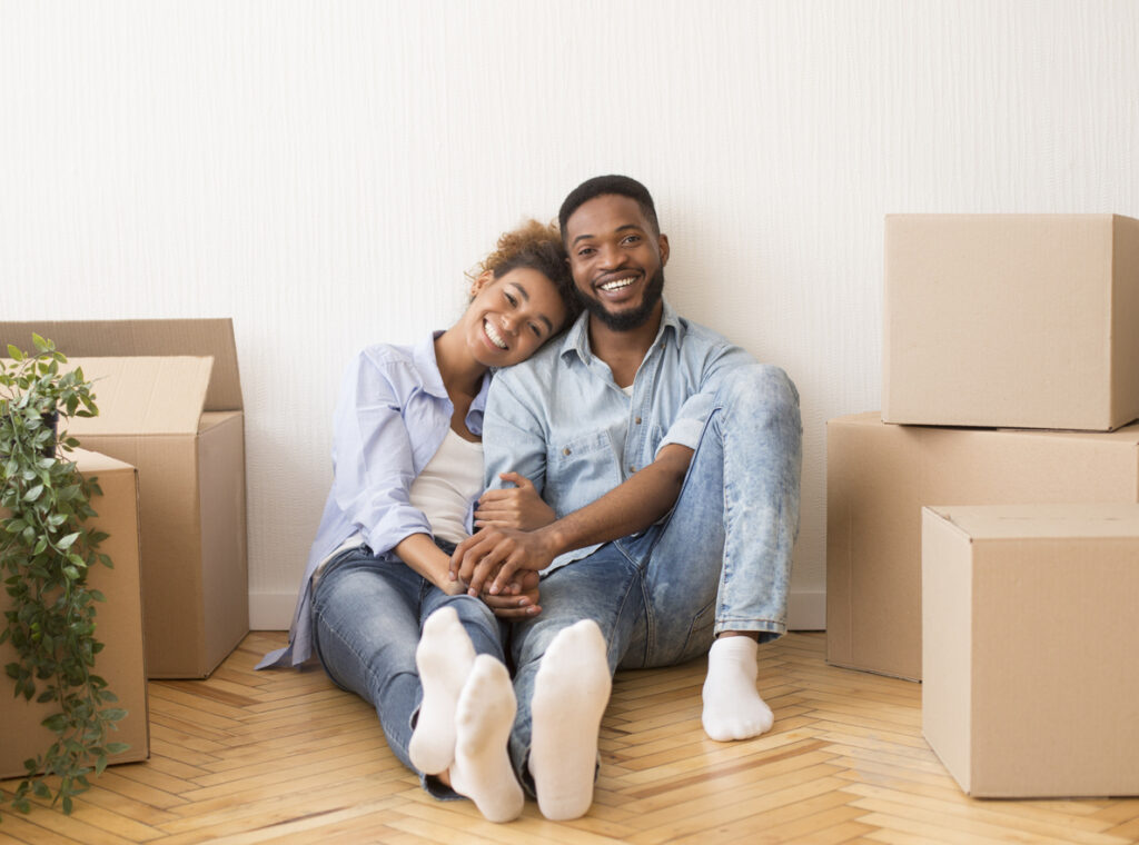 Happy African American Couple Sitting On Floor Among Moving Boxes Holding Hands And Smiling At Camera In New House.